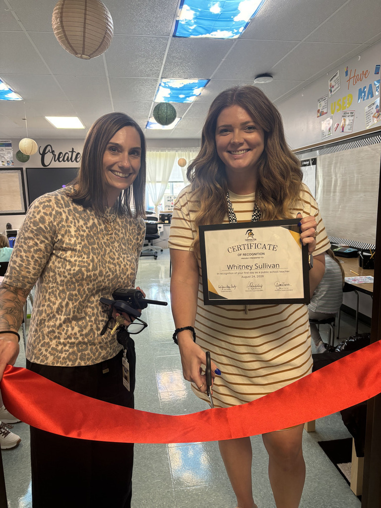 Two women stand holding a ribbon. One woman smiles and holds a certificate. The ribbon is red.