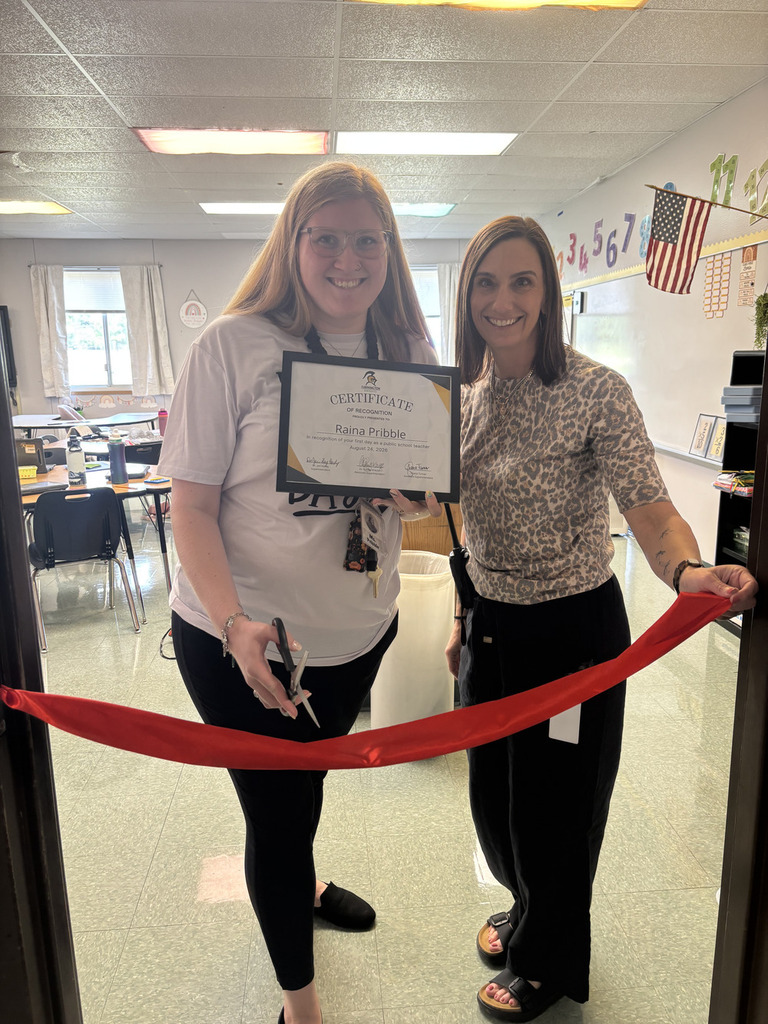 Two women stand in a classroom holding a certificate and a red ribbon. One holds scissors and a certificate.