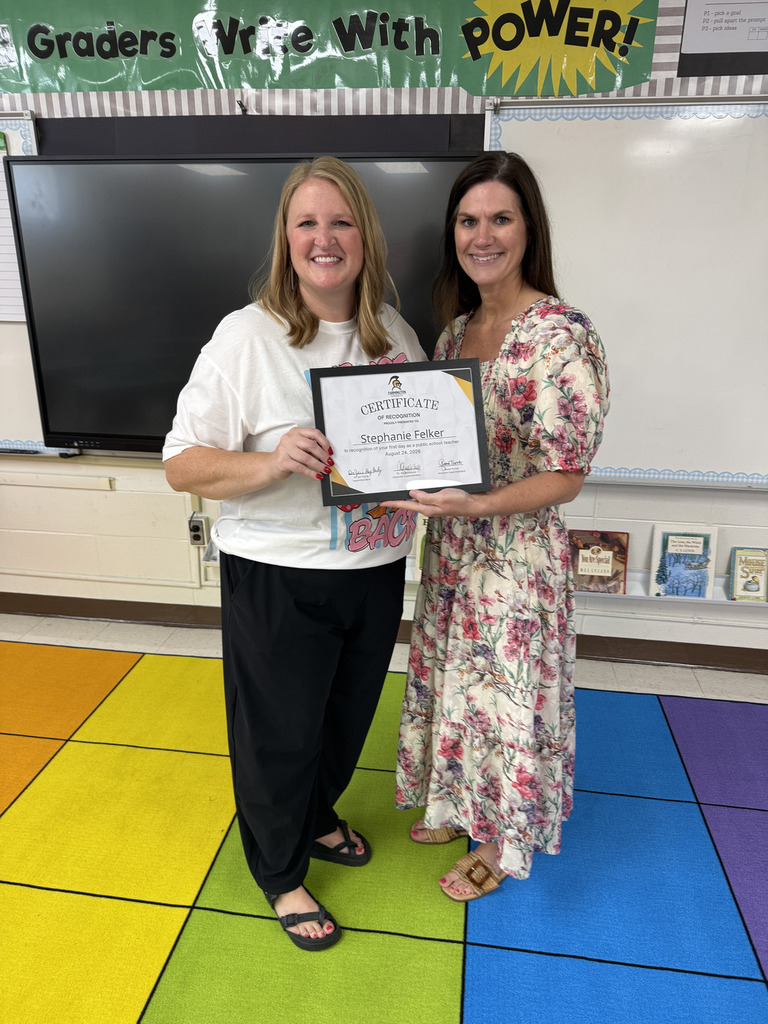 Two women stand on a colorful floor, one holding a framed certificate. They smile, with a whiteboard and books in the background.
