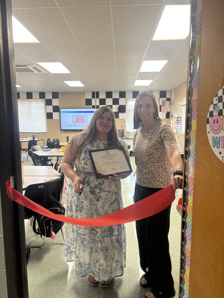 Two women in a room with a red ribbon between them. One holds a framed certificate.