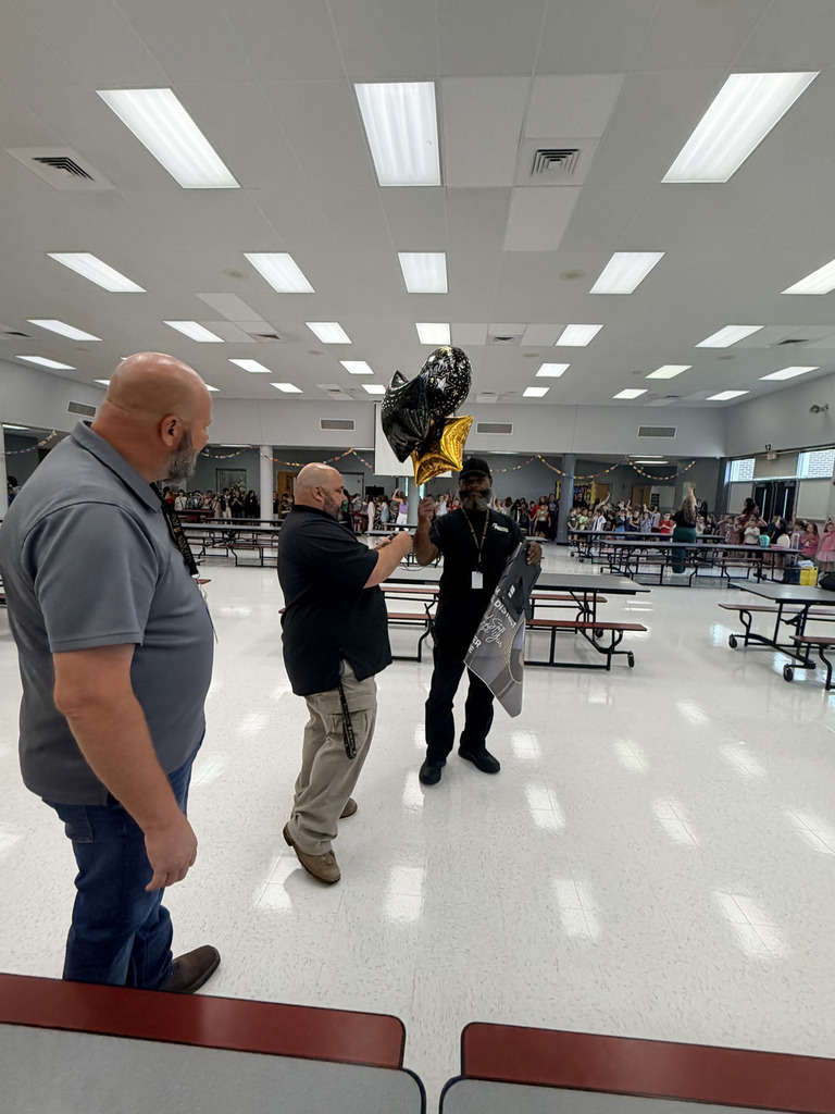 Three men stand in a cafeteria with tables and benches. One holds a trophy and a balloon.