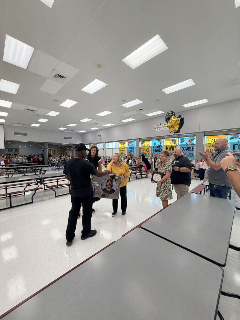 A group of people gather in a school cafeteria with tables and a stage in the background.