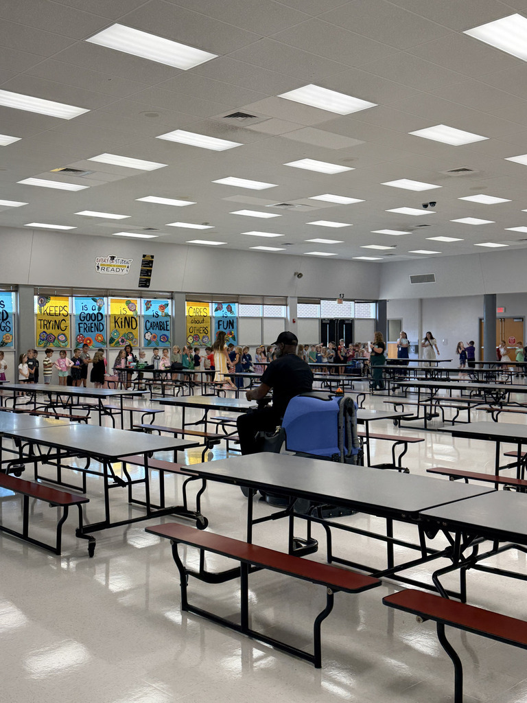 School cafeteria with many empty tables. A man sits in a chair near tables. Children in line at the far end.