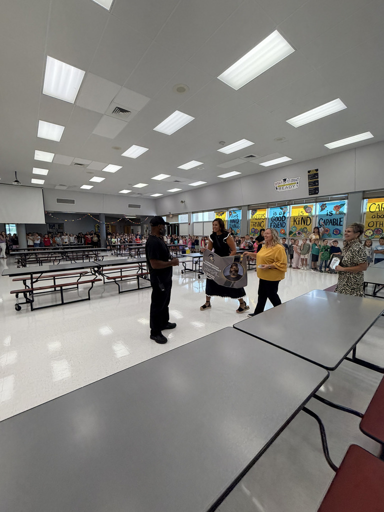 Several people stand in a room with fluorescent ceiling lights. Four adults converse at tables while others mill about.