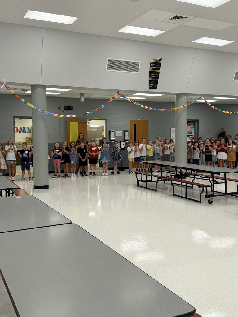 Group of children and adults in a brightly lit, spacious room with tables and a colorful banner above.