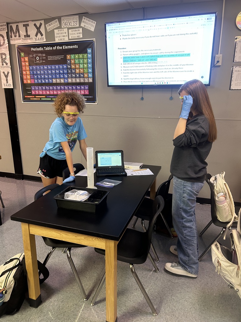 Two women, one in goggles, work at a table with a laptop and papers. A large screen is behind them.