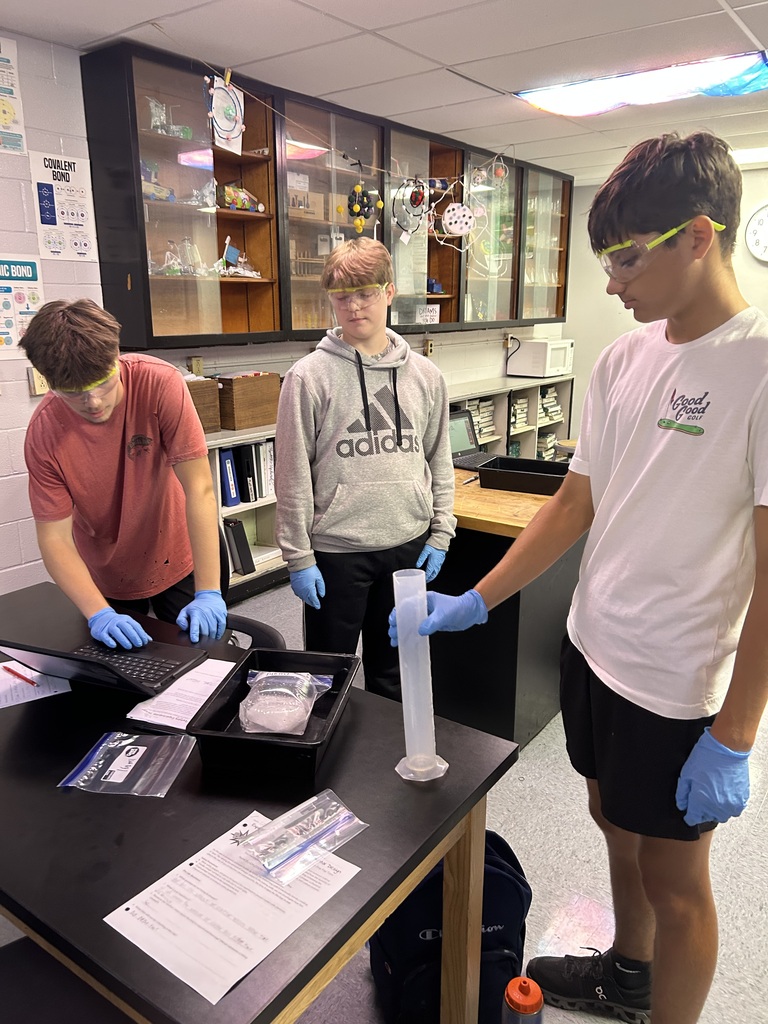 Three students in a classroom are working at a lab table with a plastic tube. One student holds the tube upright.