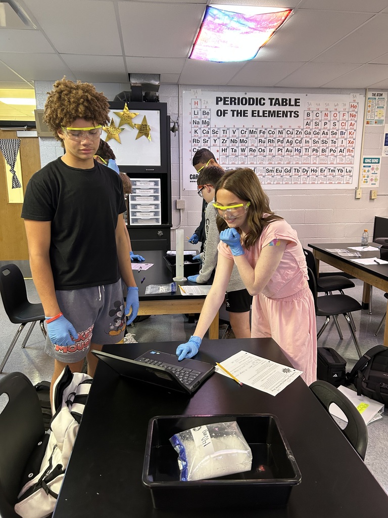 A classroom with people in lab attire. A woman in pink stands at a table with a laptop. A man in black stands behind her.