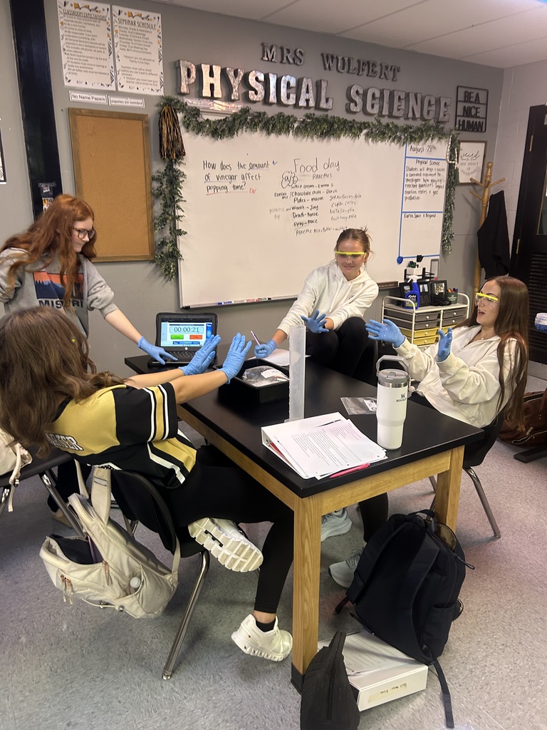 Four students wearing blue gloves, sitting around a table, in a classroom with a whiteboard and a holiday banner.