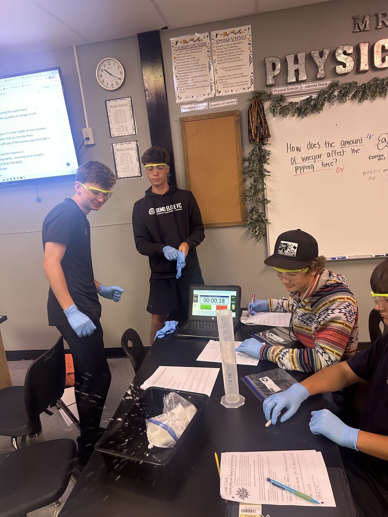 Four individuals in lab coats and gloves at a table, with a laptop, papers, and a whiteboard.