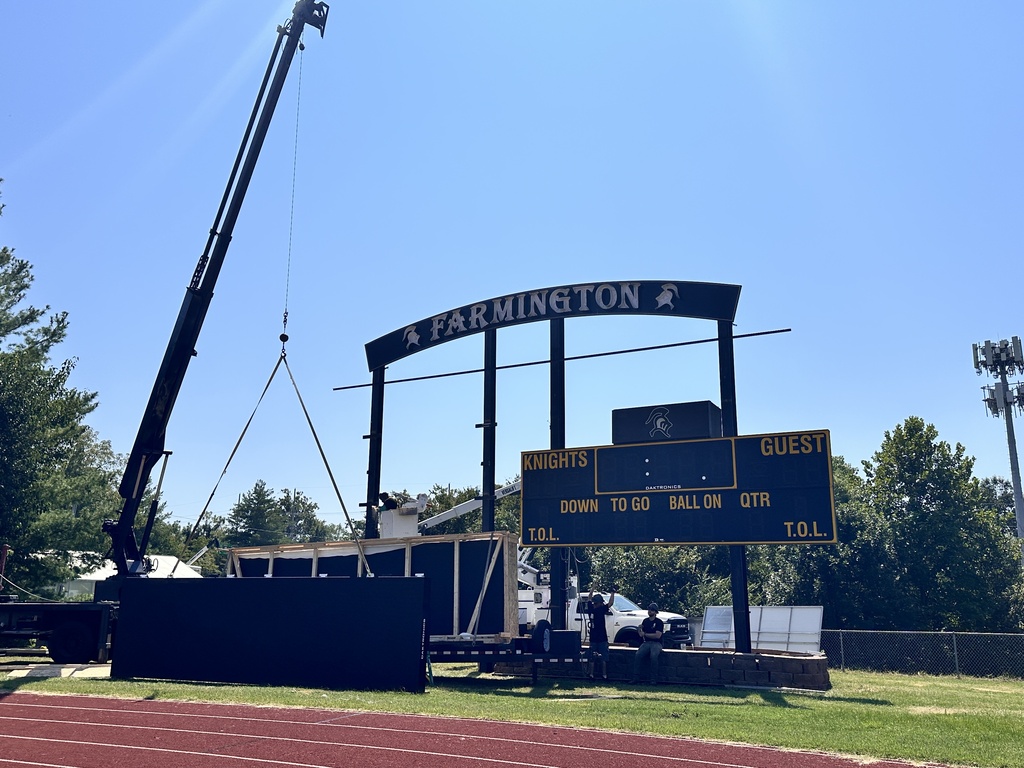 A crane lifts a scoreboard reading "Parmington" above a field with a red track. Behind it, a clear sky and trees.