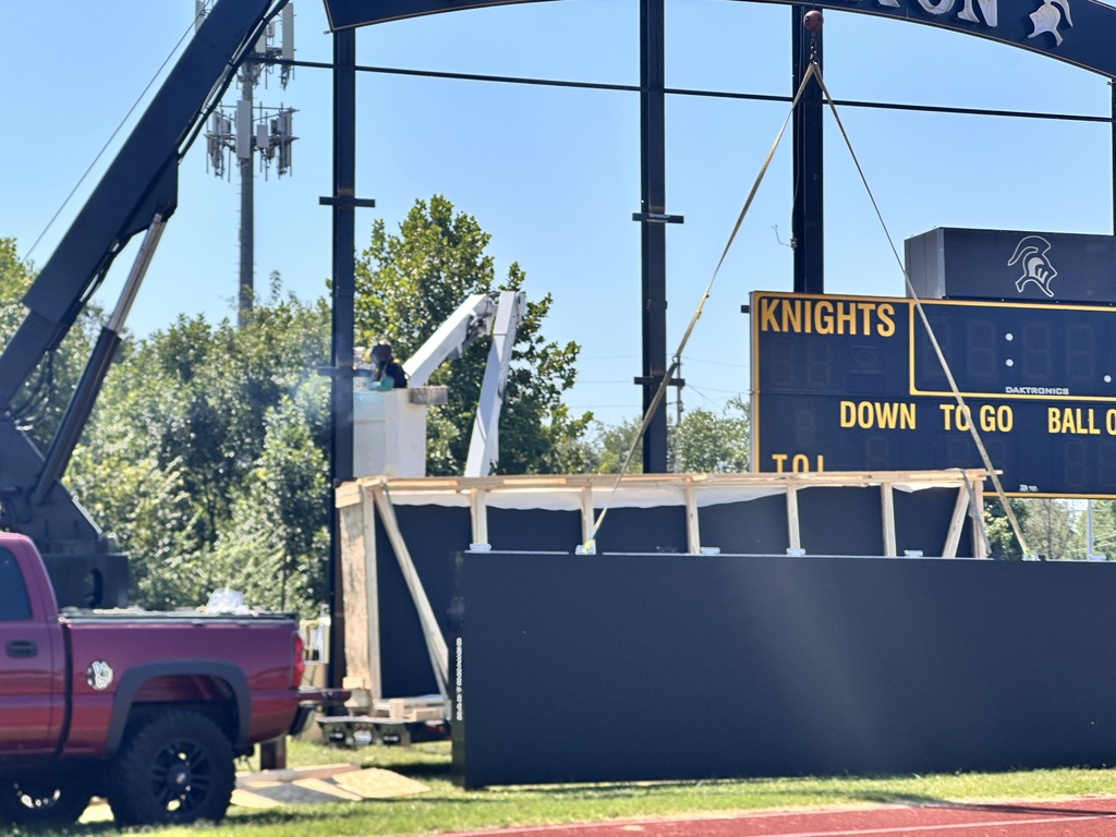 A red pickup truck beside a sports field with a large scoreboard under construction. Trees and a clear blue sky in the background.