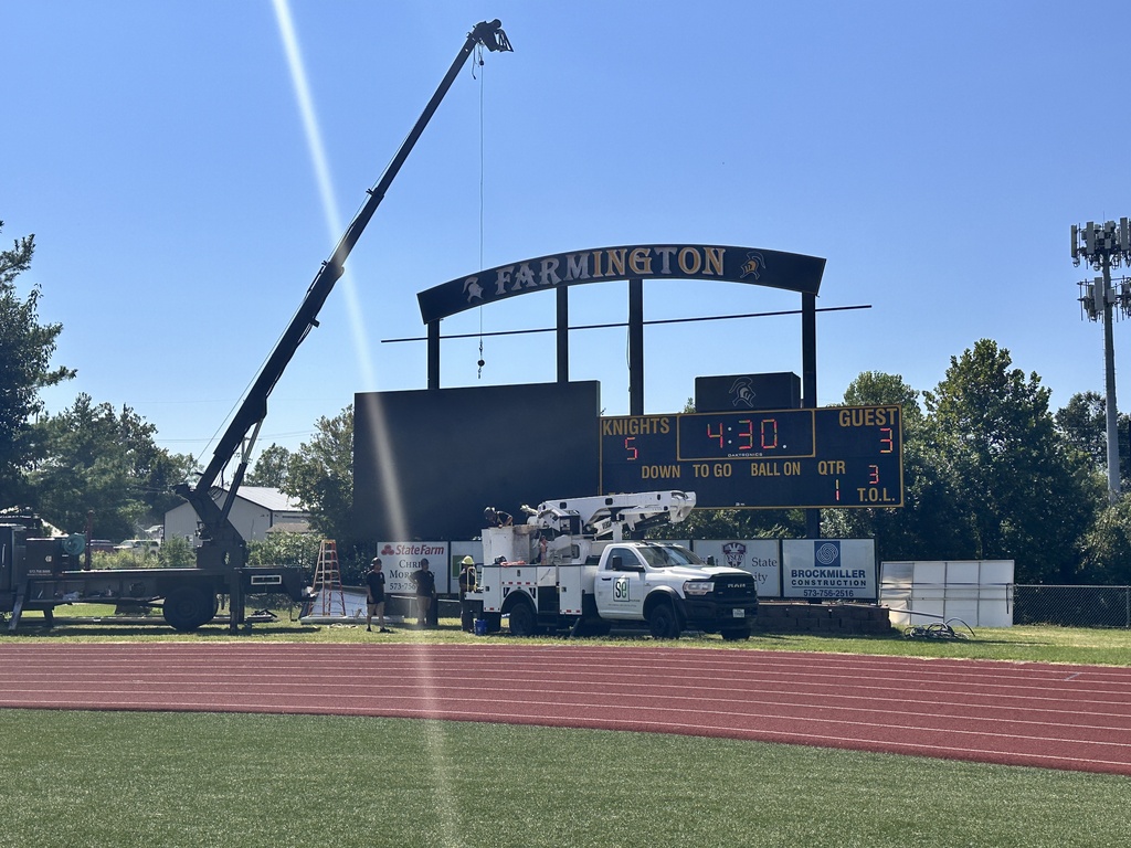 Crane next to a large scoreboard, a white vehicle parked on a field, a red track, and a grassy area.