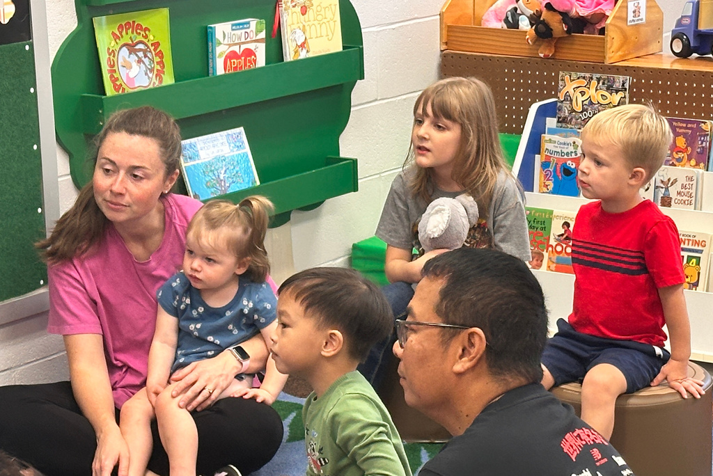 A woman and a man sit with a group of young children. They are looking at books on shelves.