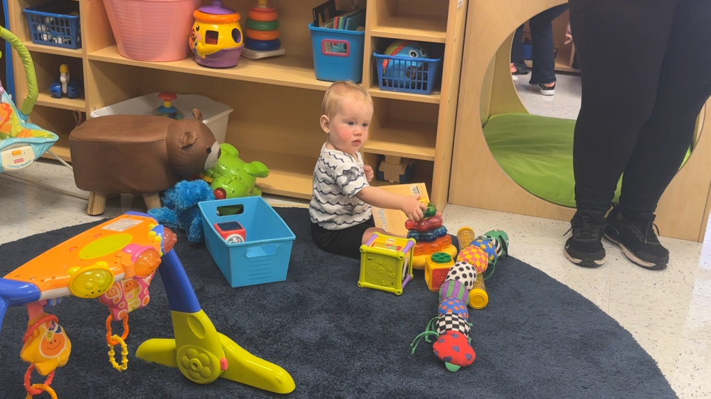 A baby in a striped shirt sits on a rug playing with toys, a wooden shelf behind them.