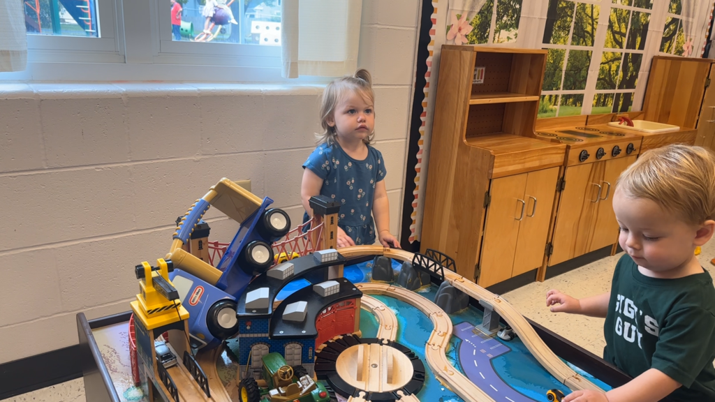 Two children, a girl and a boy, are playing with a wooden toy train set in a room.
