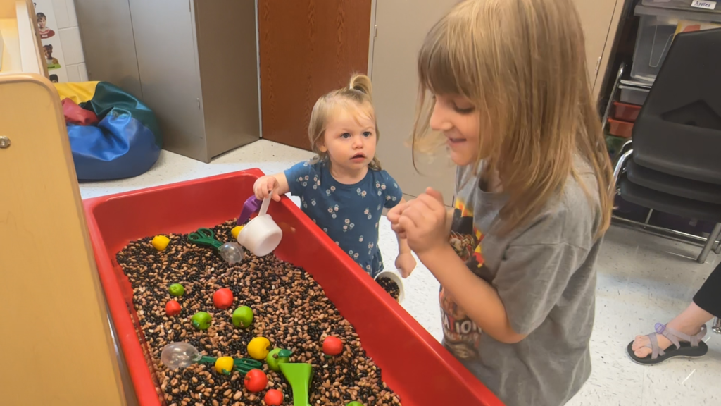 Two young girls playing with a sensory bin filled with beans and toys. One holds a cup.