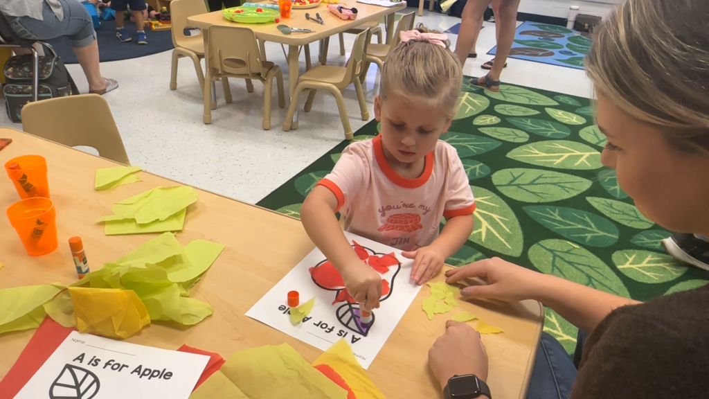 A child in a pink shirt works on an arts and crafts project while a woman assists.