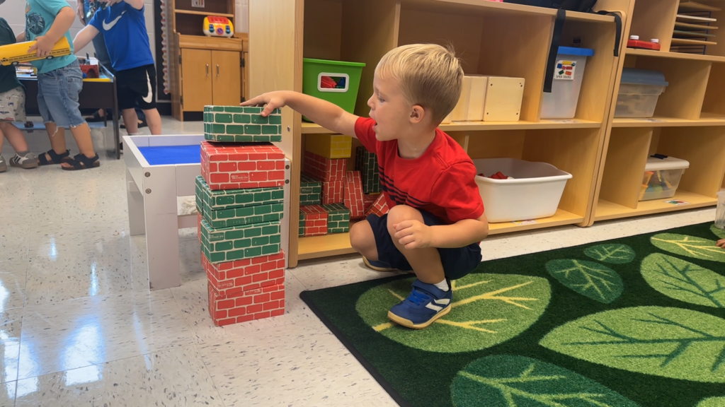 A young boy in a red shirt stacks blocks. A green rug with leaf patterns and shelves with storage boxes are in the background.