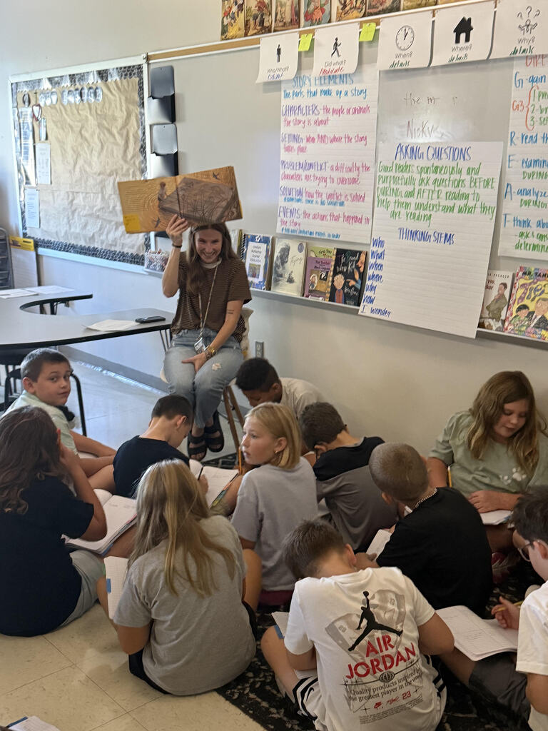 A classroom with students seated in a circle. A teacher holds a book above her head.