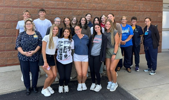 A group of people stands outside in front of a brick wall. Most wear casual clothing.