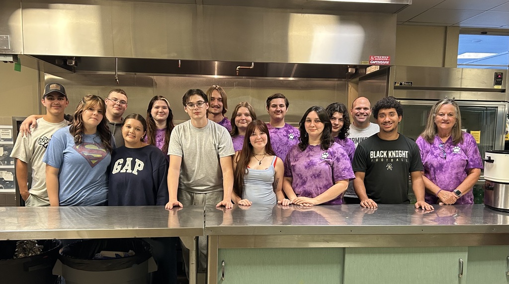 A group of people stand in a kitchen, wearing purple shirts. A countertop is in the foreground.