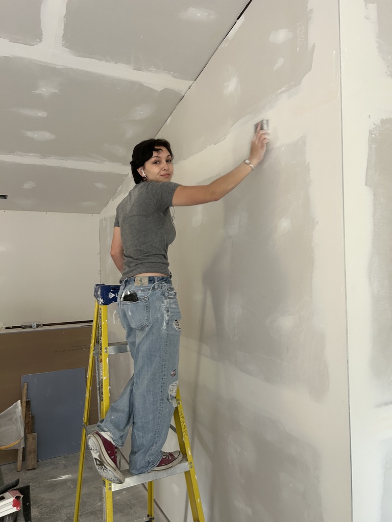 A person wearing a gray shirt and jeans stands on a yellow ladder, painting a white wall.