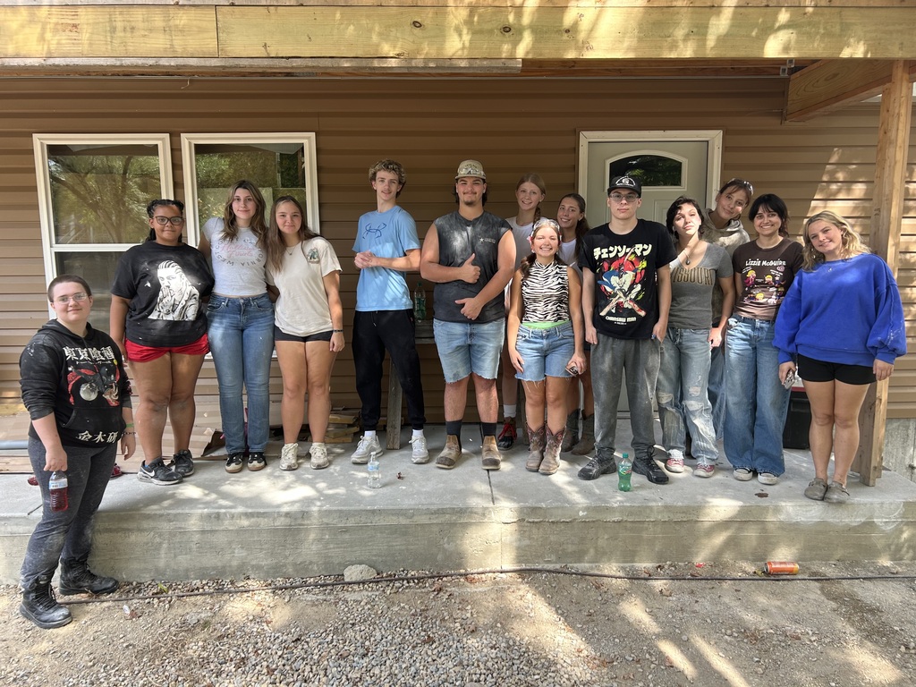 A group of people in casual attire stands in front of a house with windows and a door.