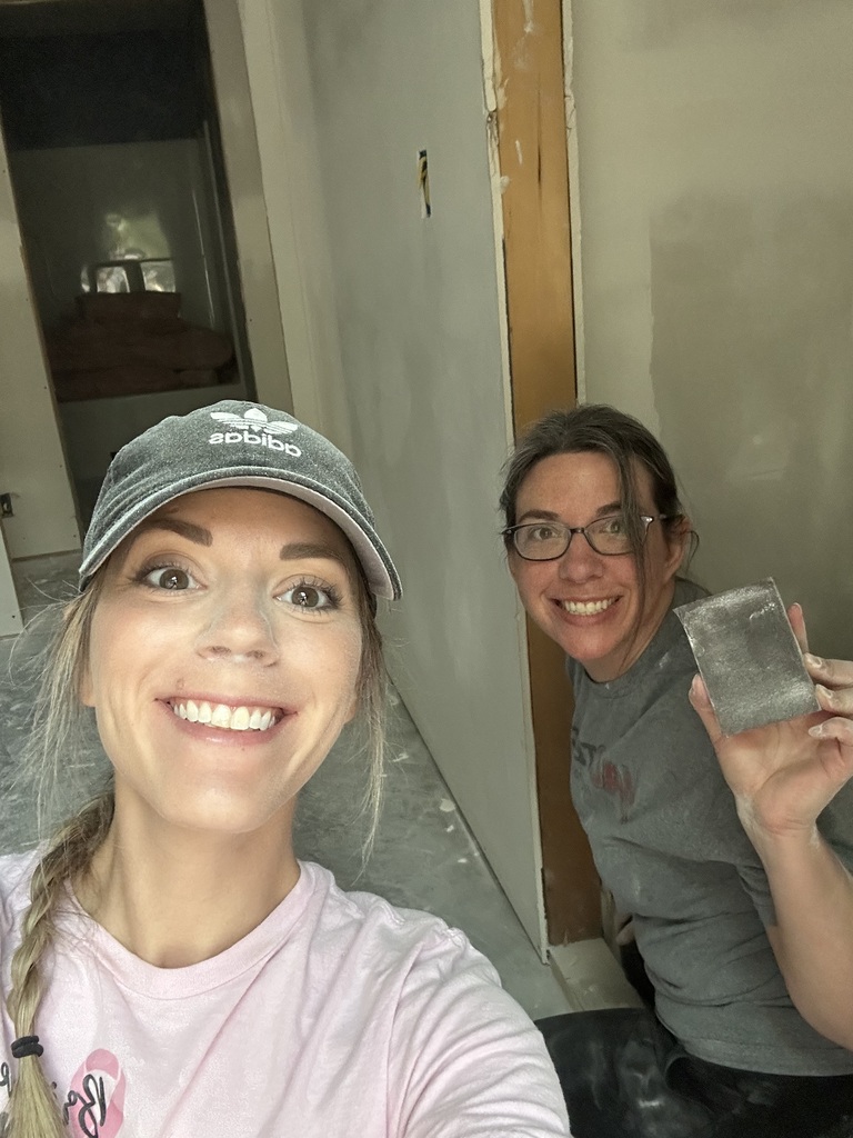 Two women are smiling, one in a pink shirt and cap, the other in a gray shirt and glasses. They are in a room with white walls.