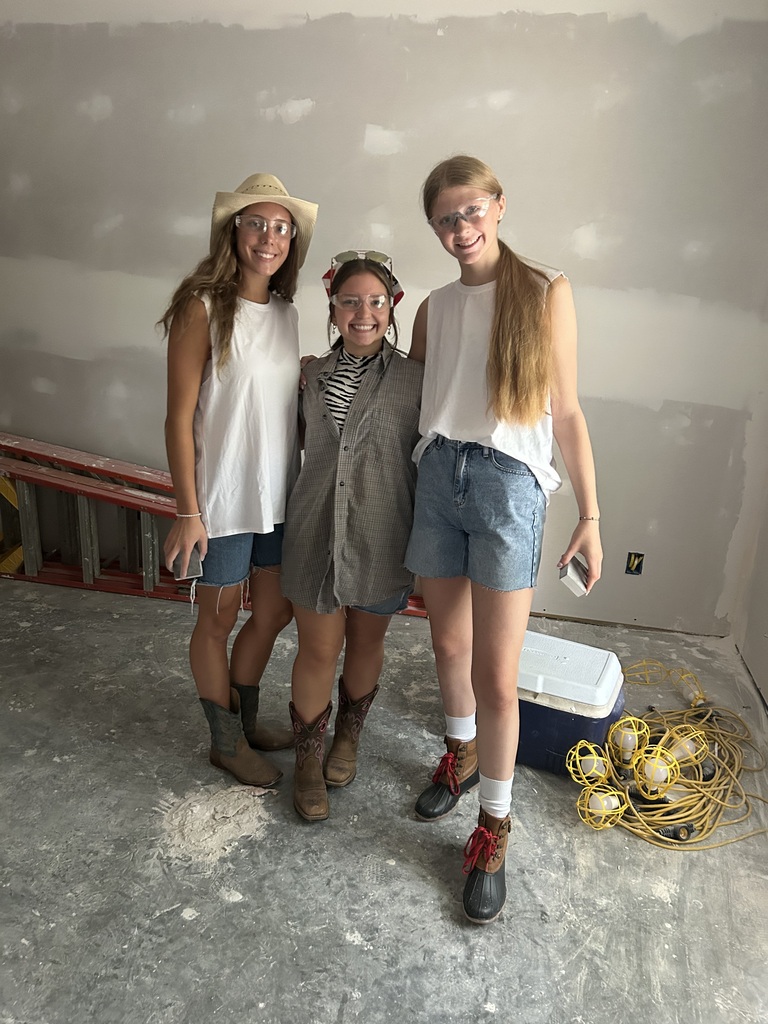 Three people stand in a room with unfinished walls. One wears a hat, another has glasses, and the third wears shorts.