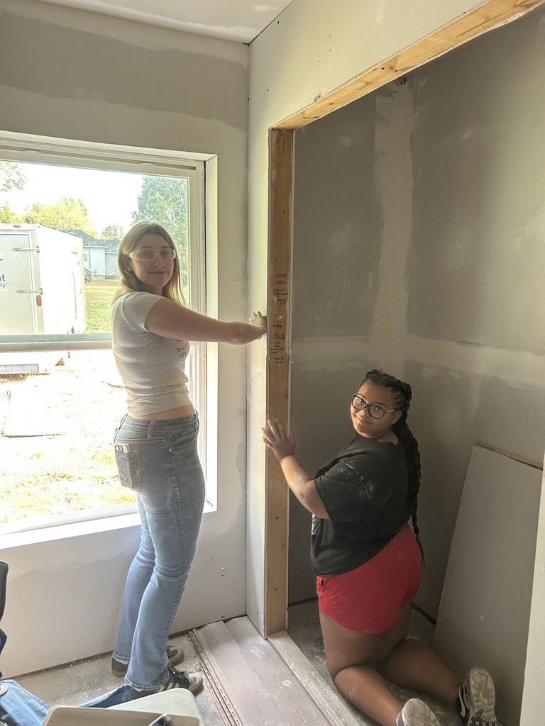 Two women work in a room with unfinished walls, one holding a door frame while the other crouches, wearing glasses. A window is to their left.