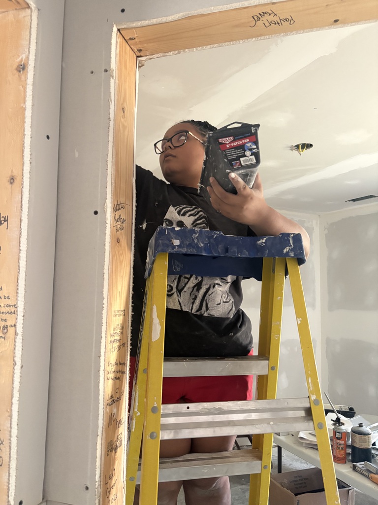 A person in glasses, a black t-shirt, and red shorts stands on a ladder holding a drill.