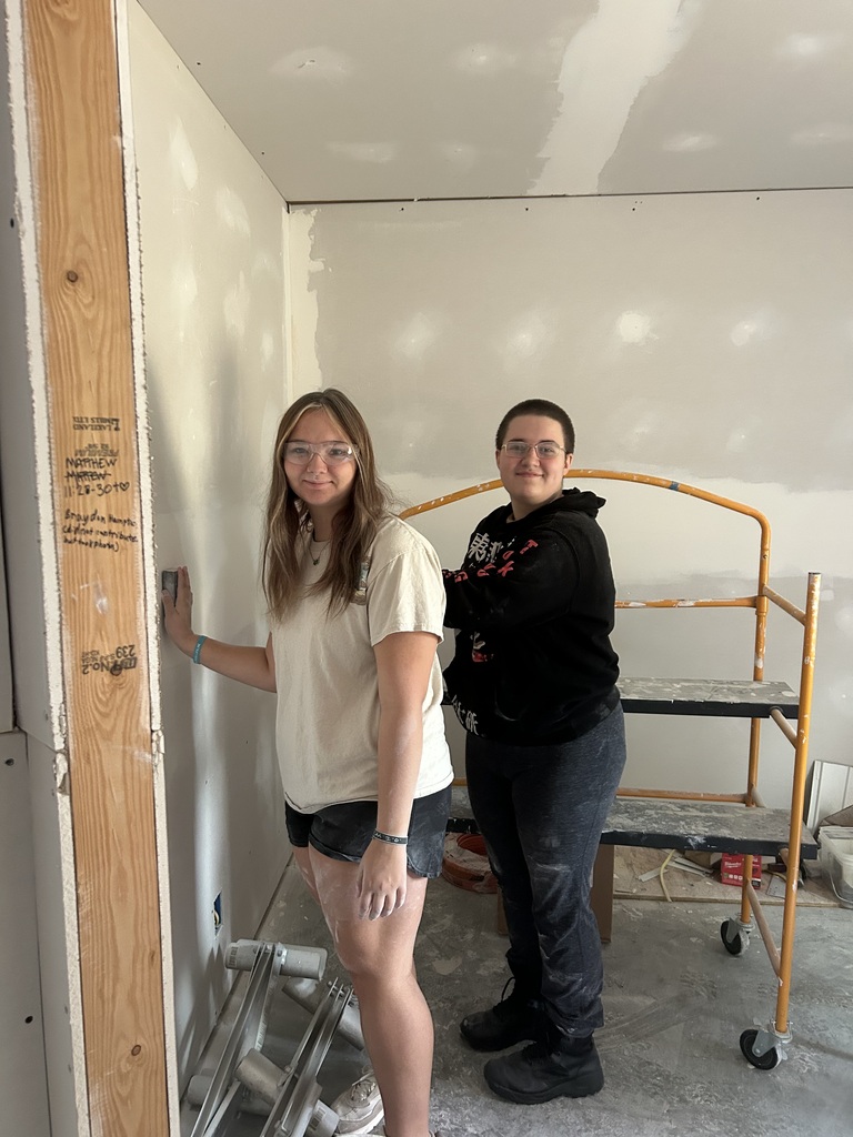 Two women, one in a white shirt and glasses, the other in a black hoodie, stand in a room under construction.