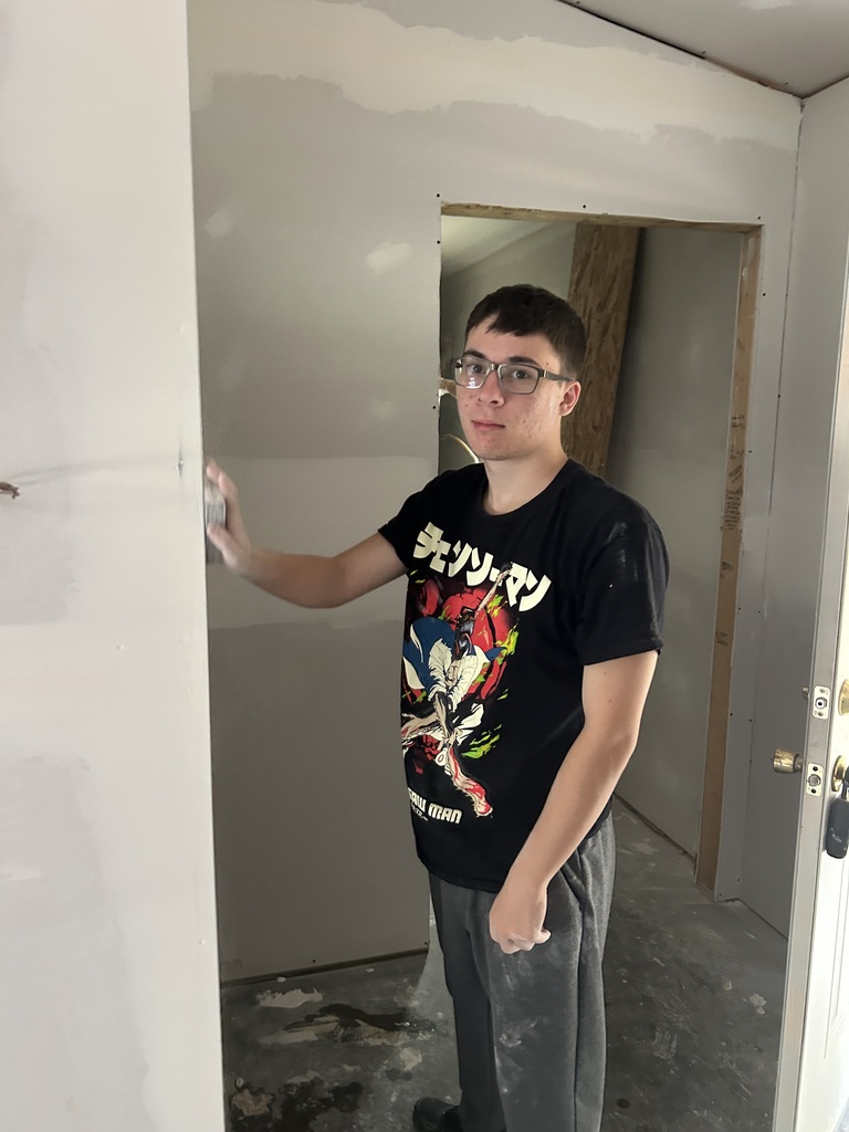 A person in a black t-shirt and glasses leans on a white wall in a room with exposed drywall.