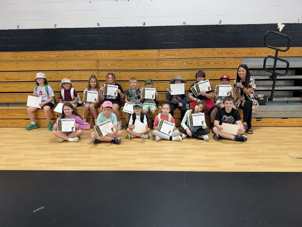 A group of children, possibly in a gym, holding certificates. They sit in a row on the floor.