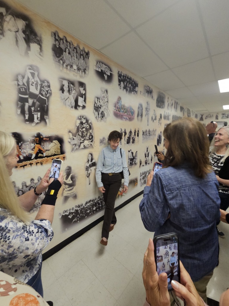 Several people stand in a hallway with a long wall covered in photographs. A man walks past them.