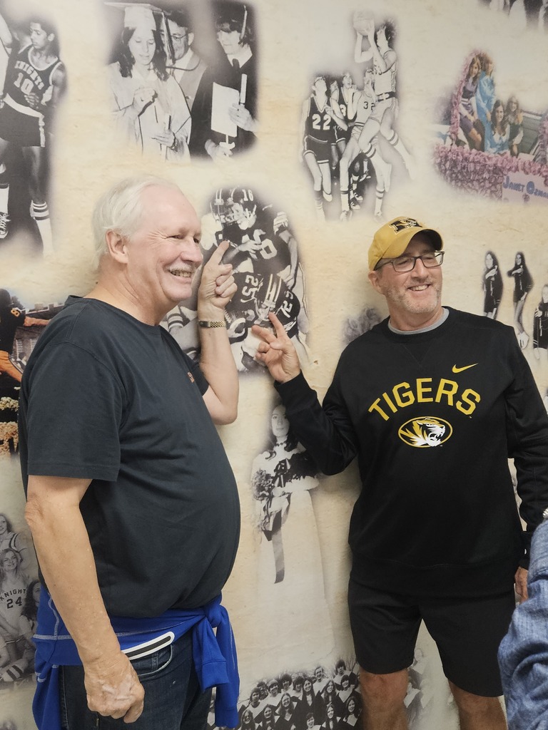 Two men stand next to each other in front of a wall with many photos of sports teams.