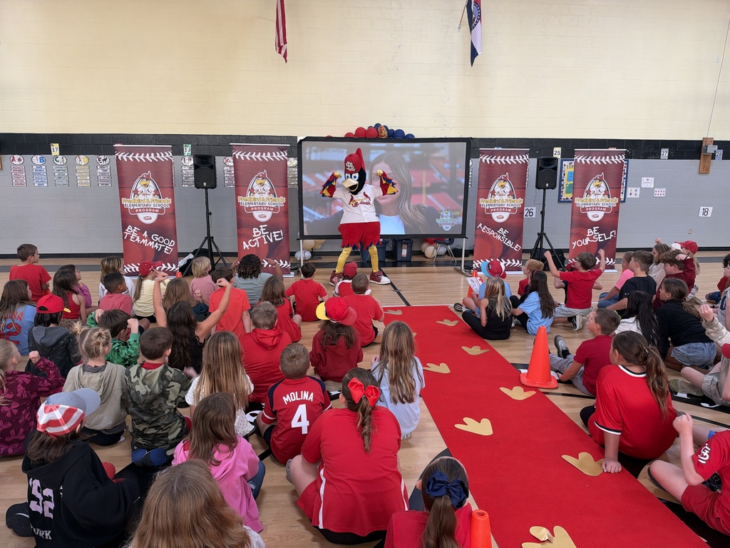 Children in red shirts sit on a floor in a school gym watching a mascot on a screen.