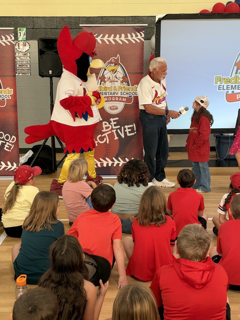 Group of children and adults watching a presentation with a mascot, a speaker, and a screen.