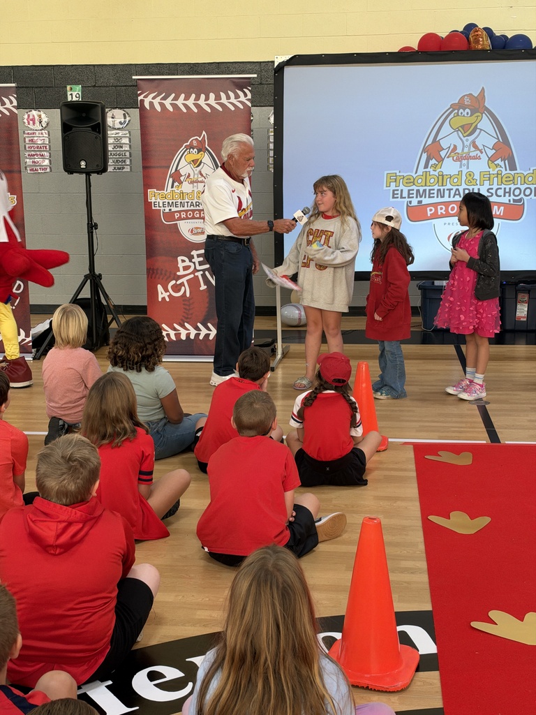 A man with a microphone speaks to children in red shirts. They sit in front of him, while others stand on a stage. Orange cones are on the floor.