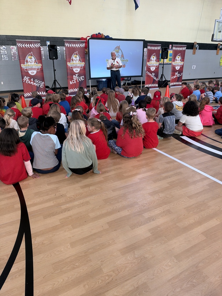 A group of children seated on a wooden floor, listening to a person speaking at a presentation with a large screen in the background.