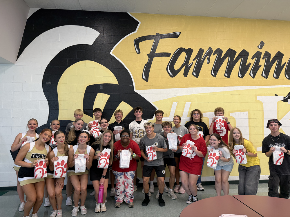 A group of students stands in front of a yellow wall with "Farmin" written on it.