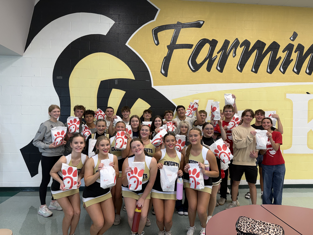 Group of cheerleaders and students standing in front of a yellow wall with "Farmington" written on it.