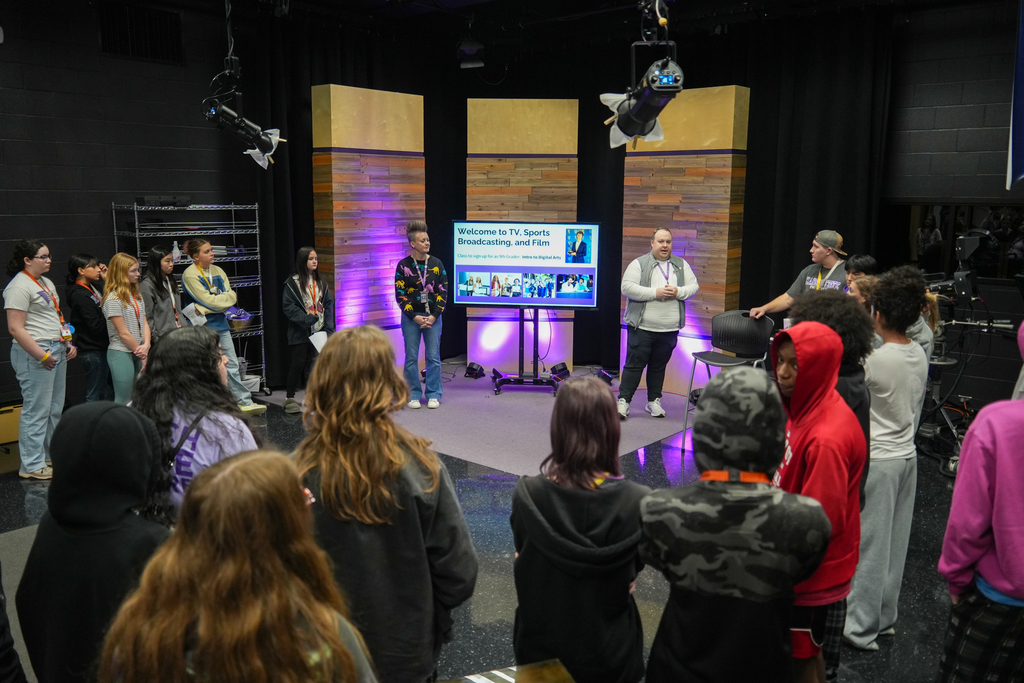 A group of people in a studio, standing in front of a stage with two individuals.