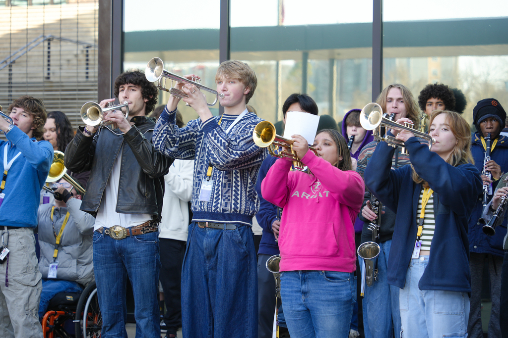 A group of people in various clothing play brass instruments in front of a building.