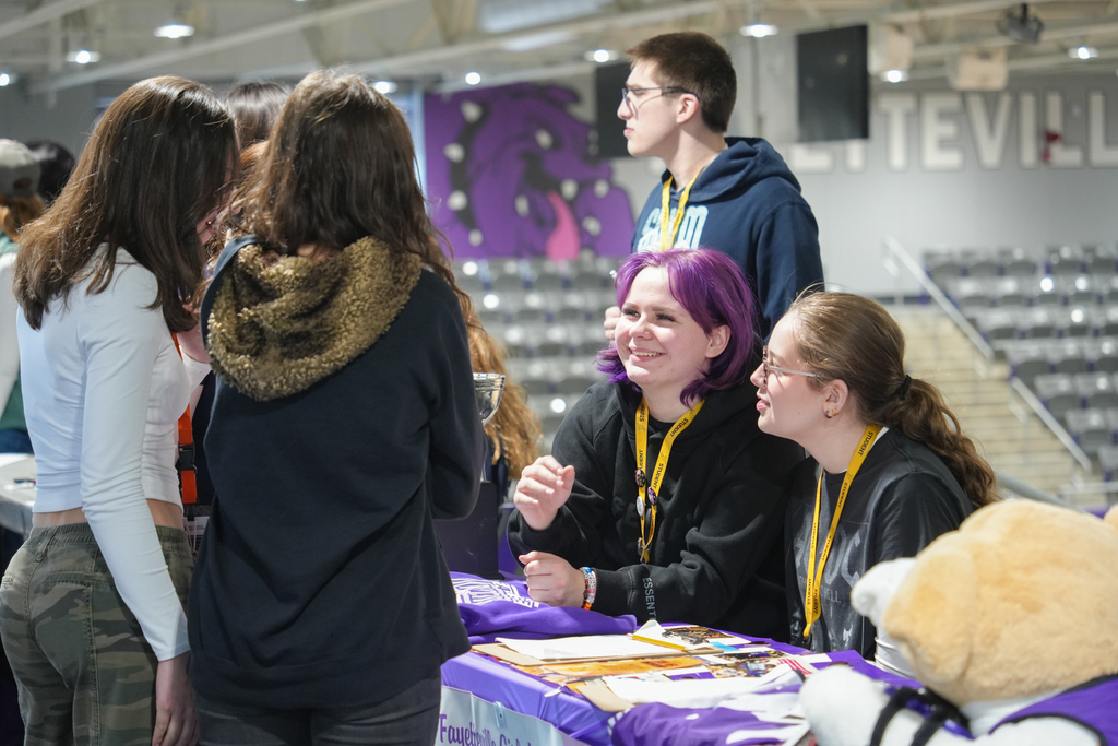 Several people interact at a table, one with purple hair, another with a lanyard. A teddy bear on the table.