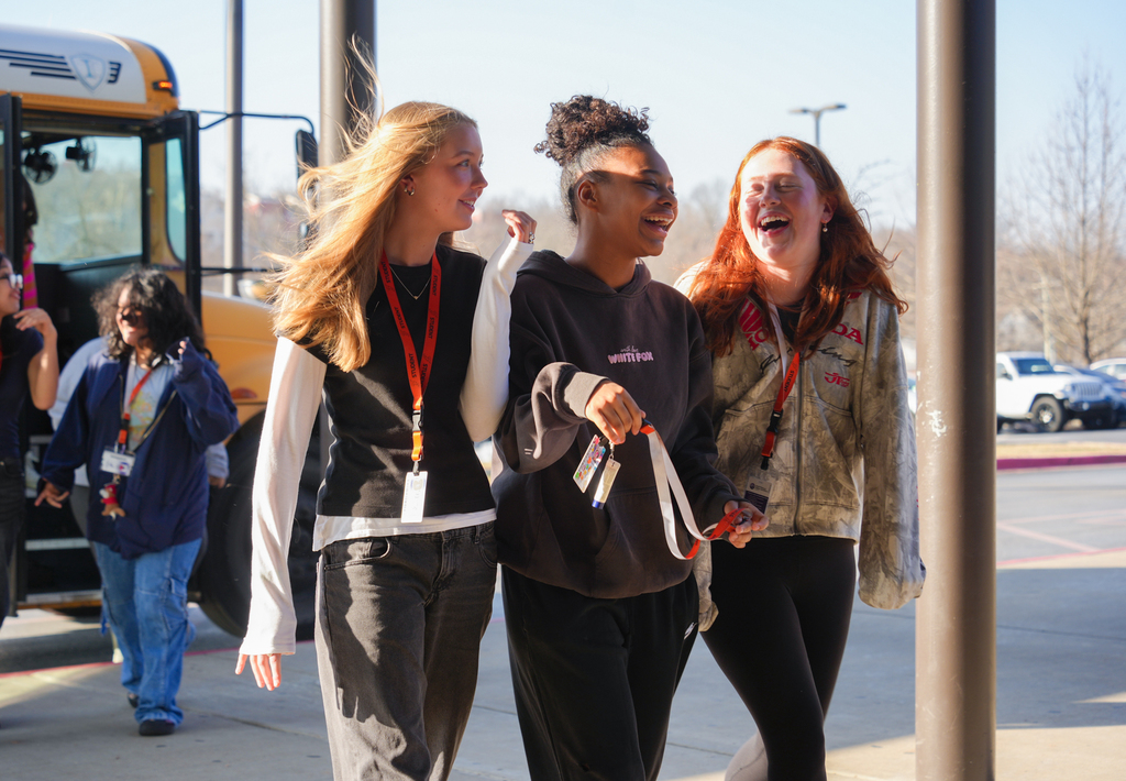 Three girls laugh while walking. A school bus stands behind them. One girl has a red lanyard.