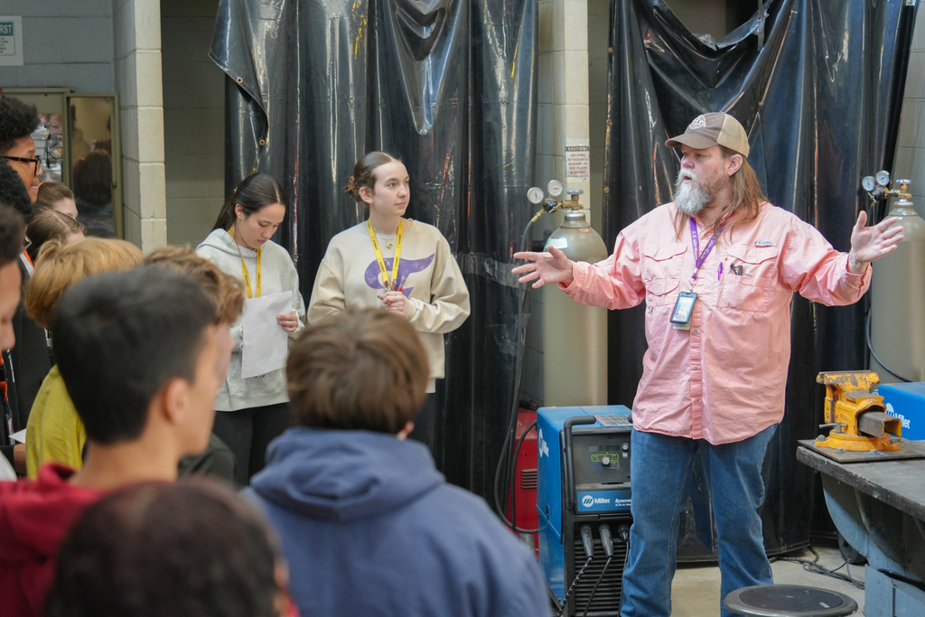 A man with a beard speaks to a group of students. Behind him, black curtains cover a wall.
