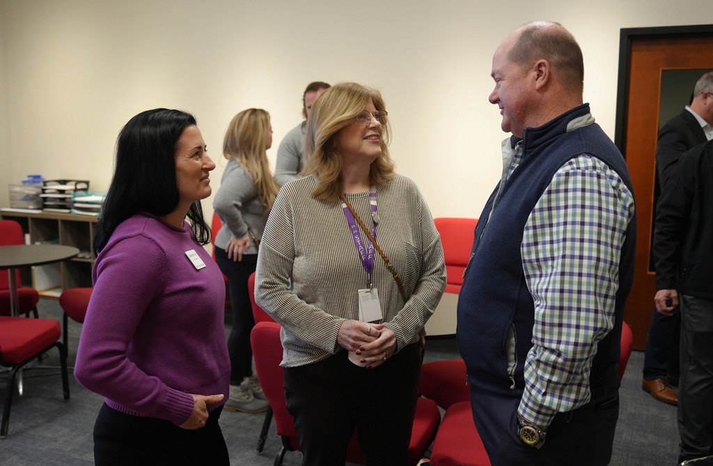 A group of adults in a room, two women in purple and gray and a man in a blue vest converse.