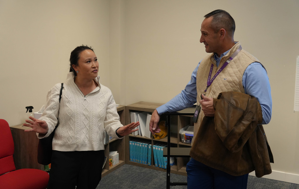 Two individuals, one with a white sweater and the other with a blue shirt, engage in conversation in an office.